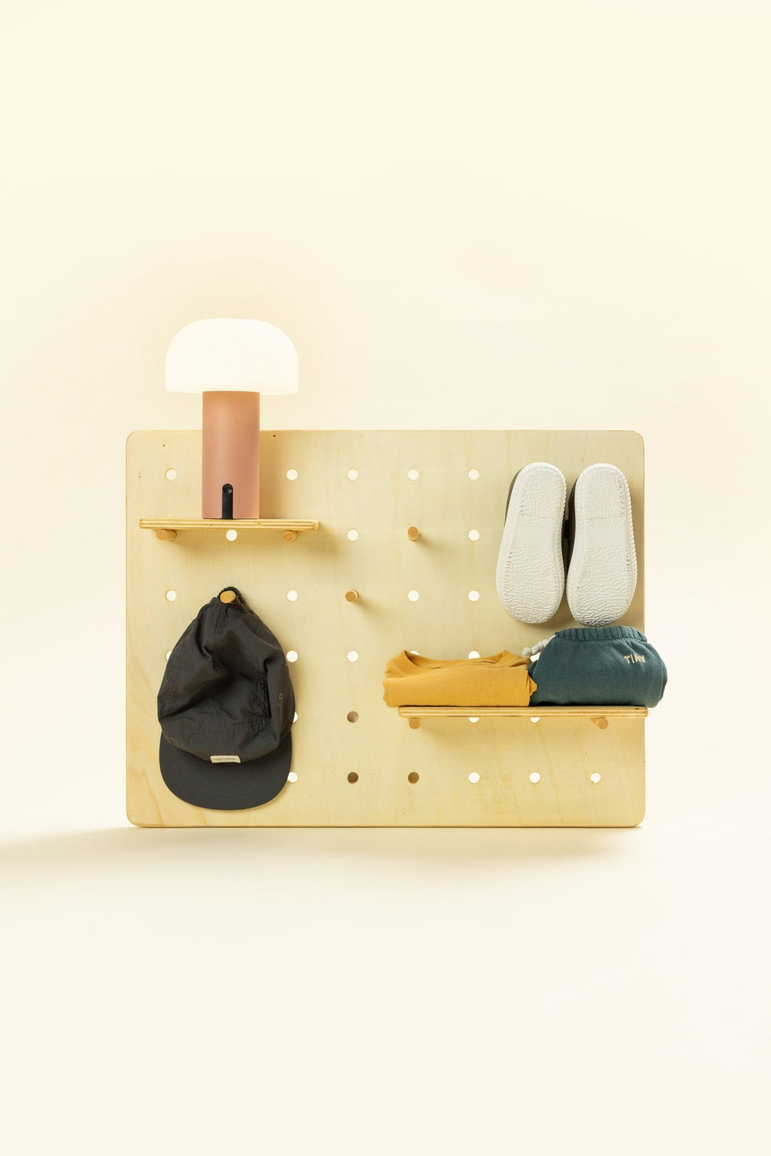 Pegboard Montessori de madera Peggy Sue de Minimaall Petit, vista frontal con zapatos, ropa infantil, lámpara y gorra colgados